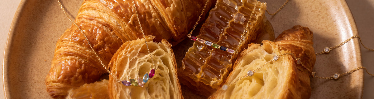 Dainty gold birthstone necklaces draped over croissant pastry and honeycomb