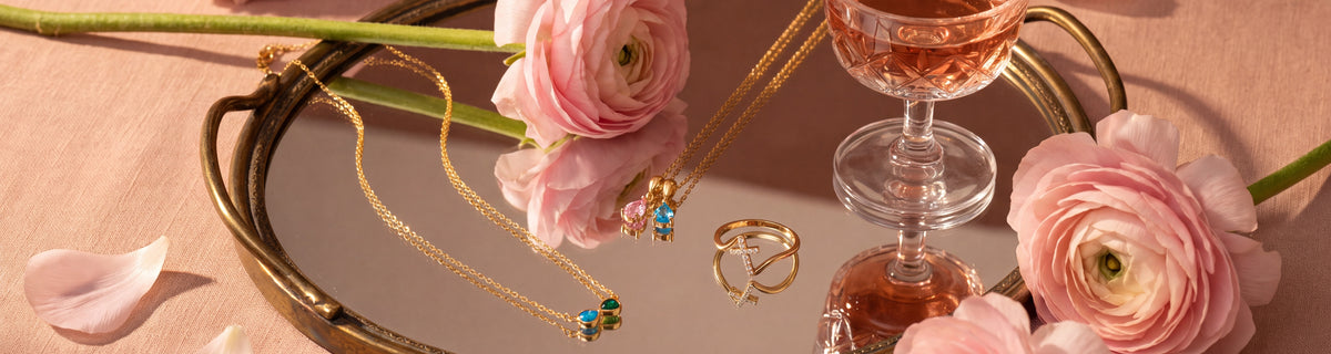 Top picks: gold birthstone necklaces and a pavé cross ring on a mirrored tray with pink ranunculus