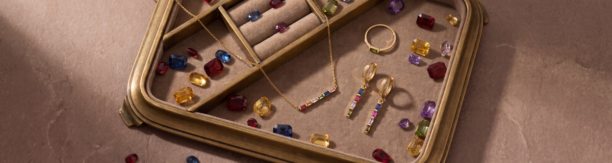 Dainty gold birthstone jewelry on a brass tray of loose coloured gemstones